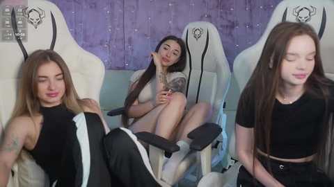 Media: Video of three young women in black outfits sitting in white gaming chairs with purple wall background. They have long hair and tattoos, appearing relaxed and casual.