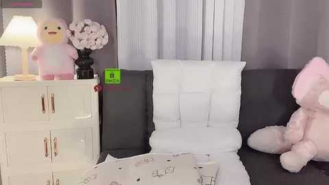 Media: A video of a cozy, minimalist bedroom featuring a plush pink bear, a white pillow, and a white cushion on a dark gray sofa. A white dresser with a lamp and floral arrangement is in the background.