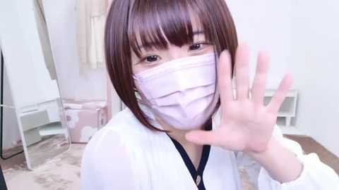 Media: Video of a young East Asian woman with straight brown hair, wearing a white face mask, black top, and white jacket, waving with her right hand, in a softly lit, minimalistic room with beige carpet.