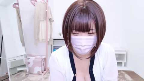 Media: Video of an East Asian woman with straight, shoulder-length brown hair, wearing a white face mask, black blouse, and white cardigan, sitting on a beige carpet in a minimalistic, white-walled room with clothes hanging and a laundry basket visible in the background.