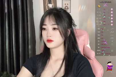 Media: Video of a young Asian woman with long black hair, fair skin, and red lipstick, sitting in a pink gaming chair. She wears a black top and has a slight pout. Background includes gaming monitors and a black curtain.