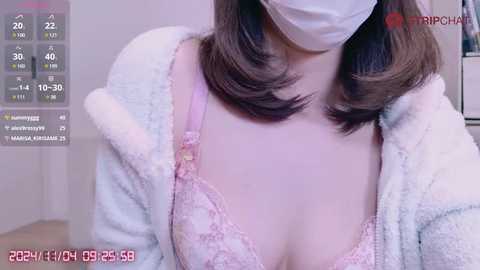 Media: Video of a fair-skinned woman with shoulder-length brown hair, wearing a white face mask, pink lace bra, and a fluffy white robe, captured indoors, with a digital thermometer display on the left.