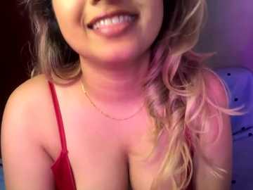 Media: Video of a smiling, light-skinned woman with wavy, blonde hair, wearing a red spaghetti strap top, revealing ample cleavage. Background is a dimly lit room with a hint of purple lighting.