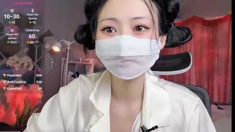 Media: Video of an East Asian woman with pale skin, black hair in buns, wearing a white medical mask, white jacket, and a black chair in a dimly lit room with red curtains.