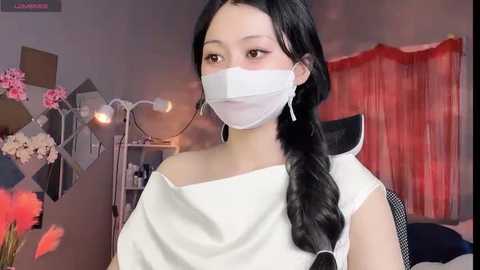 Media: Video of an East Asian woman with long black hair, wearing a white mask and white top, sitting in a dimly lit room with pink flowers and red curtains in the background.