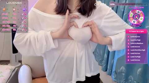 Media: Video of a young woman with pale skin and dark hair, wearing a white off-shoulder blouse, pressing her hands over her chest, against a blue curtain background. Text overlays include \"Lovense\" and \"Chat Room.\