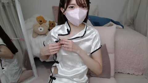 Media: Video of a young Asian woman in a white satin robe, wearing a pink surgical mask, seated in a cozy, pink-themed room with teddy bears and fluffy pillows.