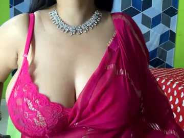 Media: Video of a woman wearing a vibrant pink saree with a lace-trimmed, low-cut blouse, revealing ample cleavage. She has a silver necklace adorned with small diamonds. Background features a colorful geometric patterned wall.