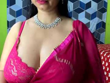 Media: Video of a South Asian woman with fair skin, wearing a vibrant pink sari with a lace bra, a silver necklace, and a geometric-patterned blue and white background.