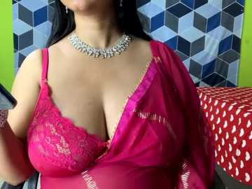 Media: Video of a South Asian woman with medium skin tone, wearing a vibrant pink lace bra and matching pink satin robe, adorned with a silver choker necklace. Background features a geometric patterned wall and a red polka-dotted bedspread.