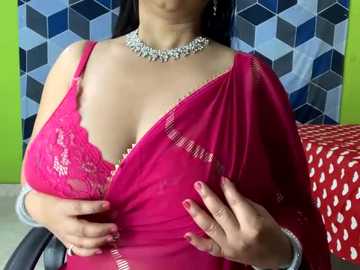 Media: Video of a South Asian woman with medium brown skin and long black hair, wearing a bright pink sari with lace detailing, a silver necklace, and a wristwatch, against a green wall with a hexagonal pattern.