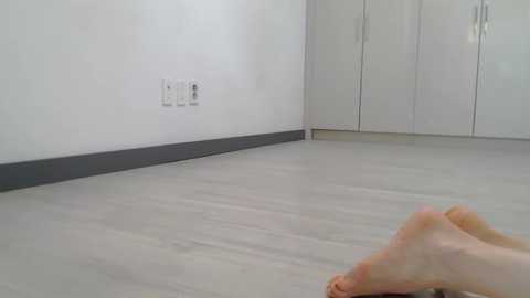 Media: Video of a minimalist, bright room with a white wall, light grey floor, and a pair of bare feet lying on the floor.