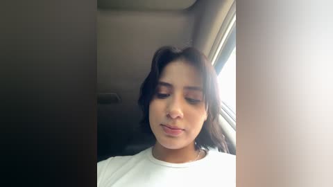 Media: Video of a young woman with medium-brown skin and straight, dark hair, wearing a white shirt, sitting in a dimly lit car. She has a serene expression and looks out the window.