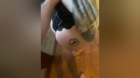 Media: Video of a pregnant woman in a black lace bra, revealing a belly tattoo of a flower and a crescent moon. She stands in a dimly lit room with wooden flooring, partially covered by a green shirt.