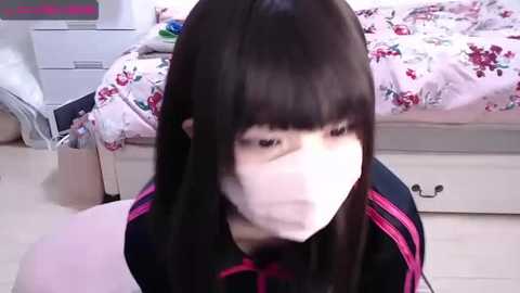 Media: Video of a young Asian woman with long black hair and a white surgical mask, sitting on a wooden floor in a modest bedroom with a pink floral bedspread and white dresser.