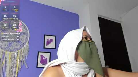 Media: A video captures a person wearing a white hooded cloak with a green face mask, standing in front of a purple wall with dreamcatcher art and framed purple artwork.