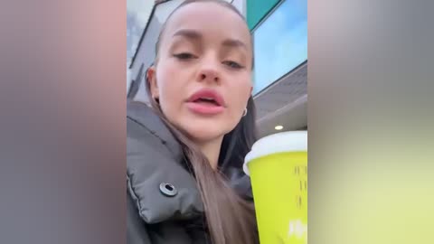 Media: Video of a young woman with light skin and long brown hair, wearing a black jacket, holding a bright yellow coffee cup with a white lid. Background shows a blurry cityscape.