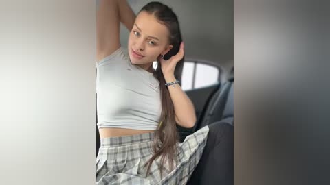 Media: Video of a young woman with long brown hair, wearing a white crop top and plaid skirt, sitting in a car, smiling.