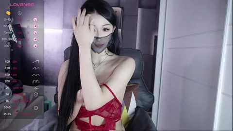 Media: Video of a young Asian woman with long black hair, wearing a revealing red lace lingerie top, seated on a grey chair in a dimly lit room.