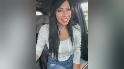 Media: Video of a smiling woman with long black hair, wearing glasses, a white top, and blue jeans, seated in a car.