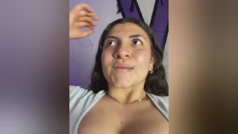 Media: A video of a Latina woman with long black hair, wearing a light gray top, showing cleavage, and a gold necklace, looking surprised with a hand raised. Purple and white wall background.