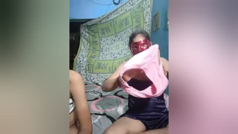 Media: Video of a young woman with light skin, wearing a red mask and black shorts, holding a pink bra, sitting on a bed with a floral-patterned sheet.