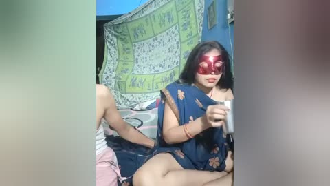 Media: Video of a woman with long black hair, wearing a red mask and blue floral dress, sitting on a bed with a green and white tapestry in the background.