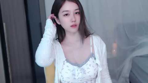 Media: Video of a young East Asian woman with light skin, straight brown hair, wearing a white floral-patterned top and cardigan, adjusting her hair in a softly lit room with white curtains and a bed.