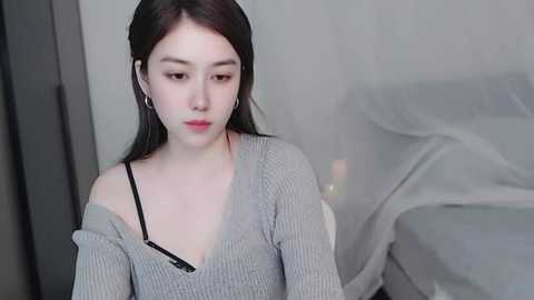 Media: Video of a young Asian woman with fair skin, straight black hair, and light makeup, wearing a gray off-shoulder sweater and black bra strap, sitting on a bed with a white canopy.