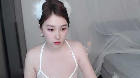 Media: Video of a young Asian woman with fair skin, dark hair, and light makeup, wearing a white lace camisole and a feathered headpiece. She is seated indoors, with a grey couch and white drapes in the background.