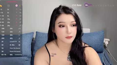 Media: A video of an East Asian woman with long black hair, fair skin, and a slender physique, wearing a black spaghetti strap top, seated on a blue couch, with a gray wall background and a TV screen displaying a chat room.