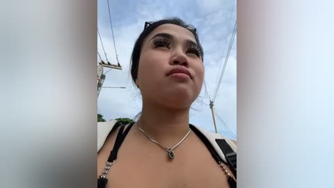 Media: Video of a young Asian woman with medium skin tone, wearing a black top, silver necklace, and dark hair. She has a neutral expression, standing outdoors with a cloudy sky in the background.