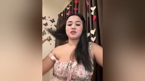 Media: Video of an Asian woman with long black hair, wearing a floral off-shoulder top, standing in a room with heart-shaped decorations on a brown curtain.