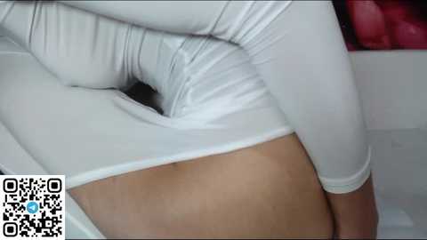 Media: Video of a woman in a tight, white, long-sleeved top, sitting with her legs spread, showing her smooth, light-brown skin. QR code in the bottom left corner.