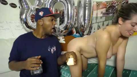 Media: Video of a muscular Black man in a navy baseball cap and shirt, holding a glass, and a nude, light-skinned woman with brown hair, leaning on a couch with a green blanket, surrounded by metallic \"2011\" balloons.