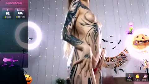 Media: Video of a nude, slender, pale-skinned woman with long blonde hair, wearing a red bracelet. Background features Halloween decorations, including a glowing pumpkin, bats, and a string of lights.