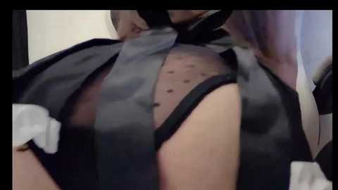 Media: A video of a woman wearing a sheer black mesh top with a black tie, revealing her polka-dot black bra underneath. Her skin is light, and the background is blurred.