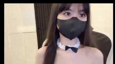 Media: Video of an Asian woman with long black hair, wearing a black face mask, black bow tie, and a white ruffled collar, seated indoors.