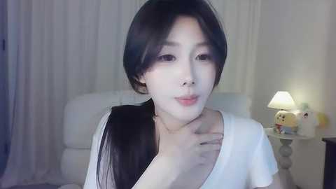 Media: Video of an Asian woman with long black hair, fair skin, and a small nose, wearing a white top, touching her neck, in a dimly lit room with a white chair and a lit lamp.