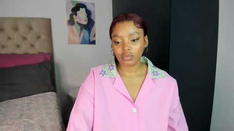 Media: Video of a young Black woman with short, red hair, wearing a pink button-up shirt with floral embroidery, standing in a modern bedroom with beige tufted headboard, colorful abstract art, and gray bedding.