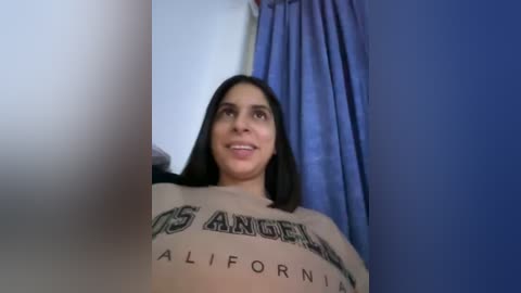 Media: Video of a smiling Latina woman with long black hair, wearing a \"Los Angeles California\" sweatshirt, standing in a room with blue curtains.