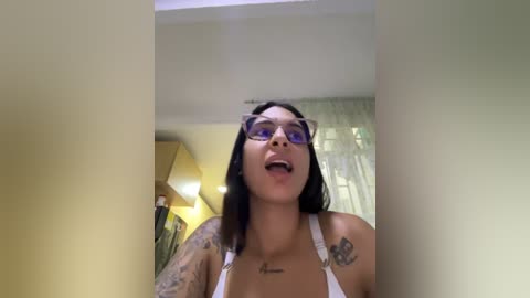 Media: A video of a young woman with dark hair, wearing purple-framed glasses and a white tank top, showcasing multiple tattoos, taken indoors with yellow walls and a window with sheer curtains.