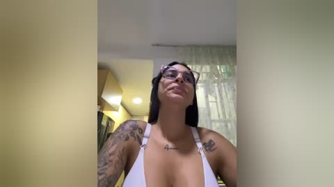 Media: Video of a tattooed woman with glasses, wearing a white tank top, standing in a modern room with light-colored walls and a window with sheer curtains.