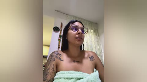 Media: A video of a young woman with medium brown skin and long black hair, wearing glasses and a light green towel, standing in a bright, softly lit room with white walls and a window.