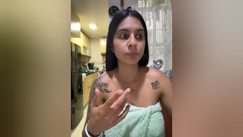 Media: Video of a young woman with medium skin tone, straight black hair, wearing a light green towel, with tattoos on her shoulders, standing in a modern kitchen with beige walls and white cabinets.