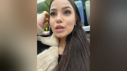 Media: A video of a young woman with long, straight black hair, wearing a beige fur coat and a black top, applying lipstick. The background shows blurred airport baggage and luggage carts.