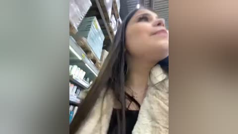 Media: Video of a smiling Latina woman with long, dark hair, wearing a beige cardigan and black top, browsing shelves in a pharmacy with green and white packaging.