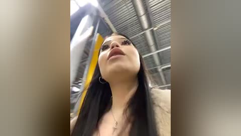 Media: A video of a young woman with long, dark hair, wearing a light brown jacket, taken from a low angle, showing her upturned face. The background features industrial metal structures and yellow accents.