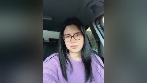 Media: Video of a young woman with long black hair, wearing red-framed glasses, a purple sweater, and a neutral expression, seated in a car with gray interior.