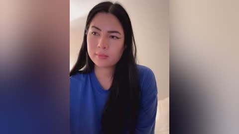 Media: Video of a young Asian woman with long black hair, wearing a blue top, looking contemplative against a blurred, neutral background.